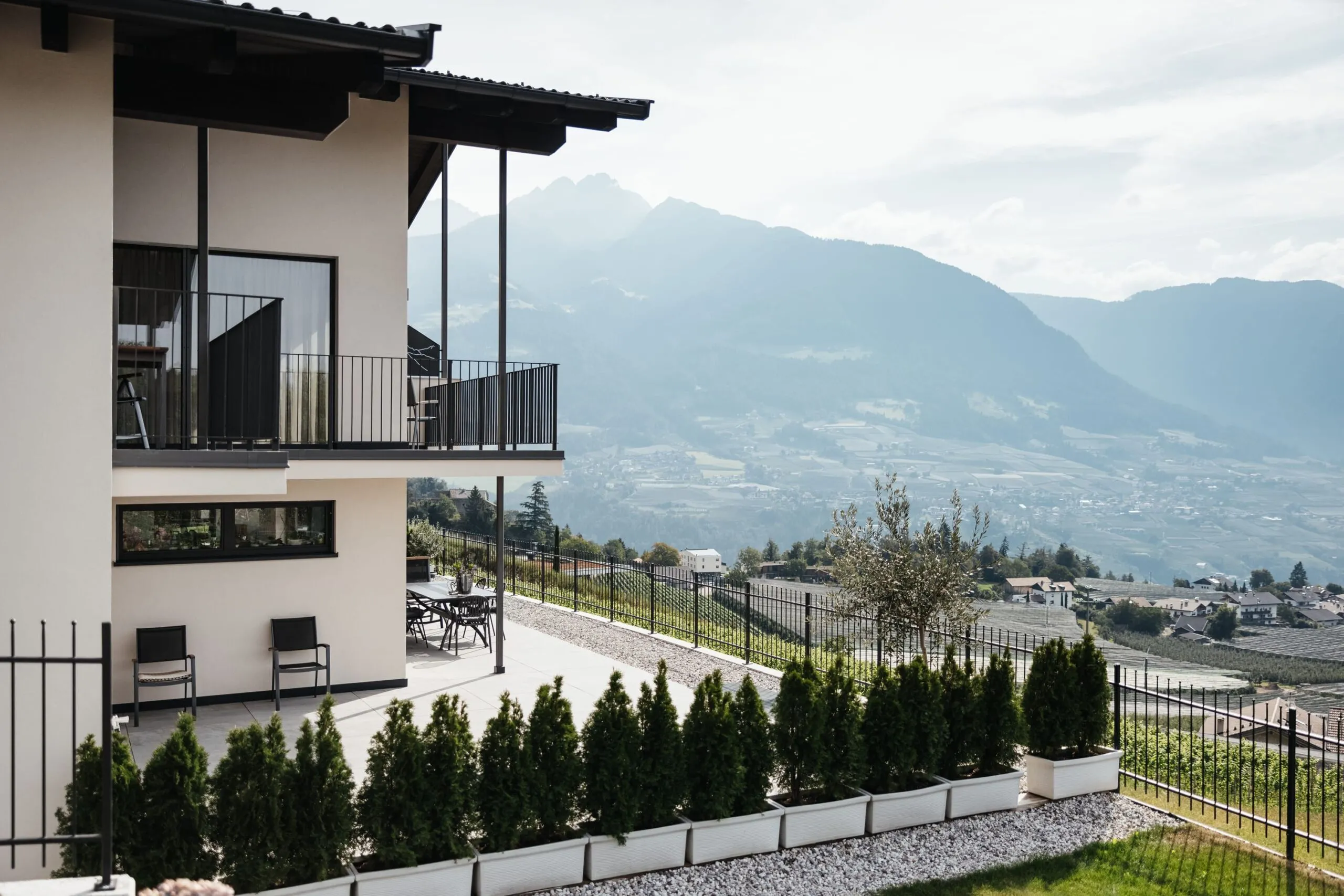 Casa moderna a livelli con balconi neri, patio con tavolo e sedie, fila di arbusti in contenitori bianchi, recinzione metallica; montagne sullo sfondo.