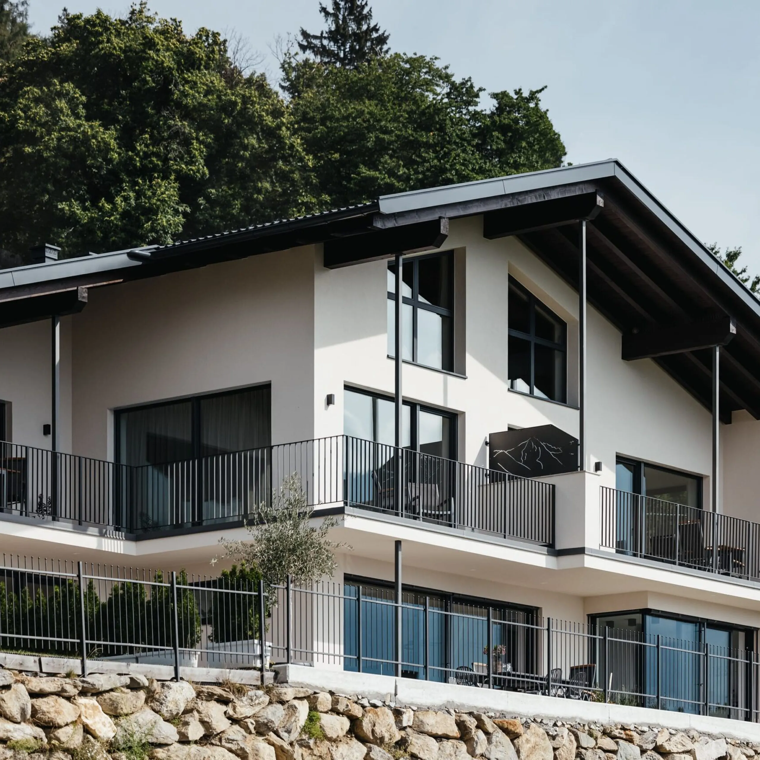Casa moderna a due piani con balconi e ringhiere in metallo, grandi finestre, su terrazzo di pietra con alberi sullo sfondo.
