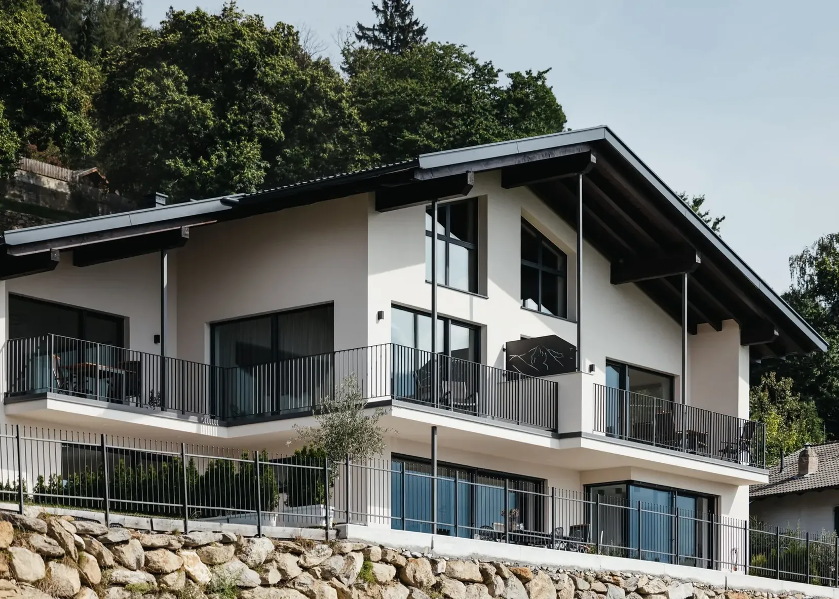 Casa moderna a due piani con balconi e ringhiere in metallo, grandi finestre, su terrazzo di pietra con alberi sullo sfondo.