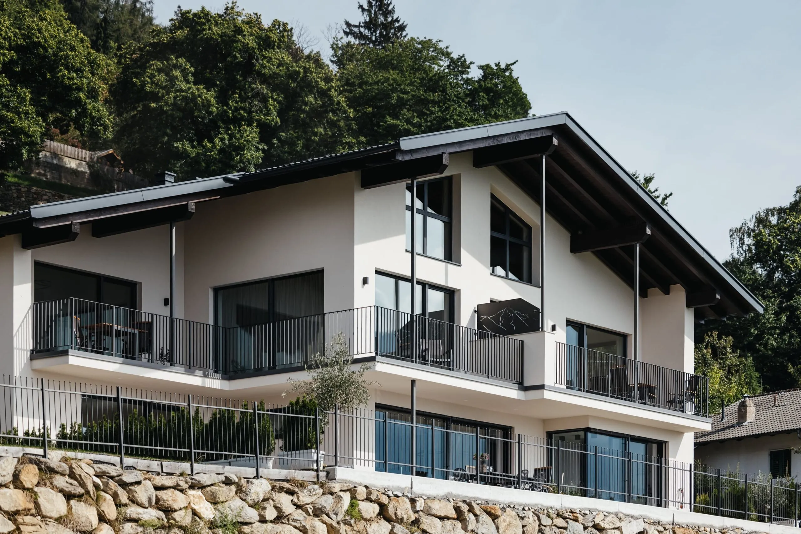 Casa moderna a due piani con balconi e ringhiere in metallo, grandi finestre, su terrazzo di pietra con alberi sullo sfondo.