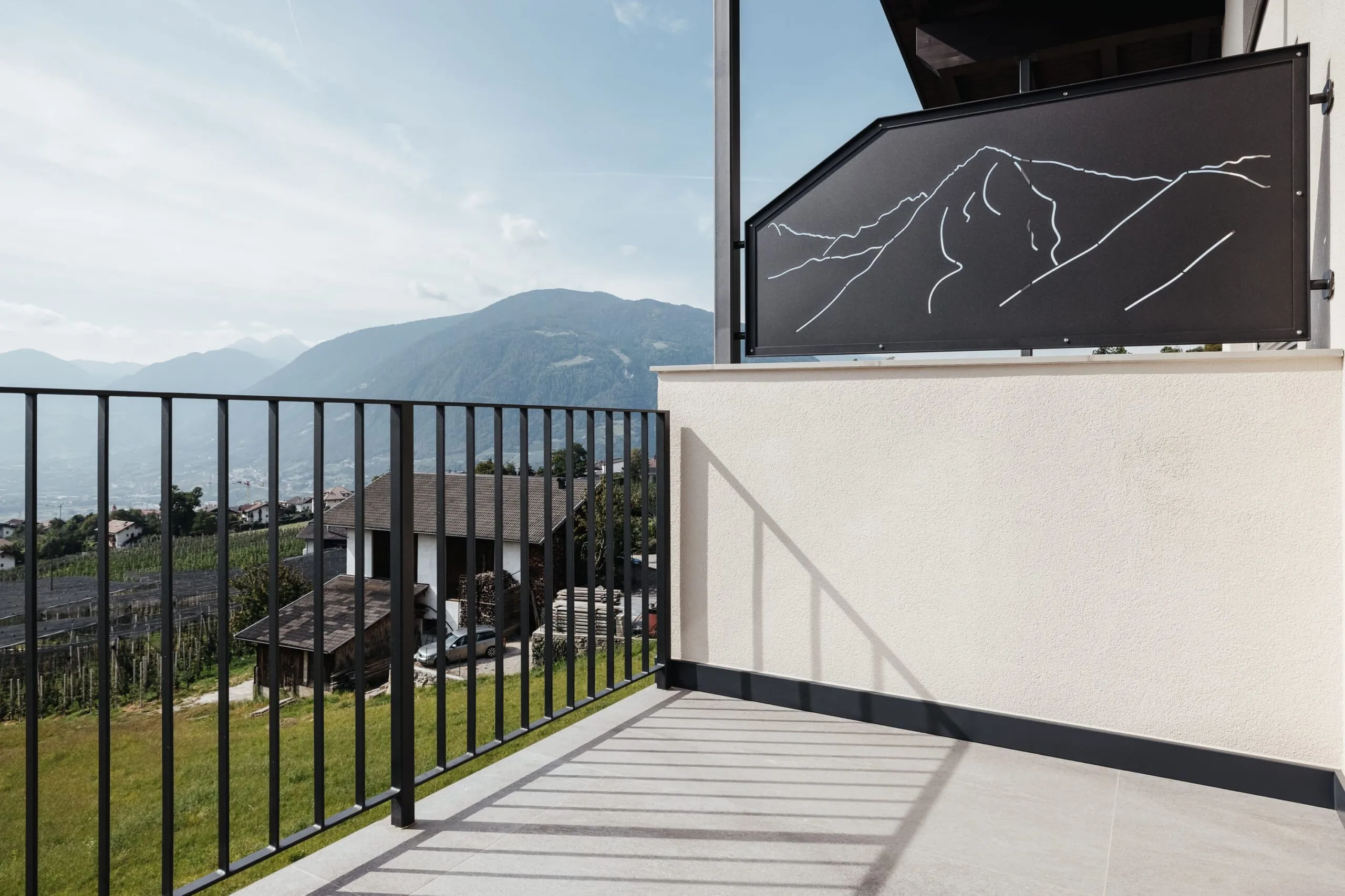 Balcone con parapetto in metallo nero, vista su villaggio e montagne; pannello nero con linee bianche che delineano montagne, fissato su parete beige.