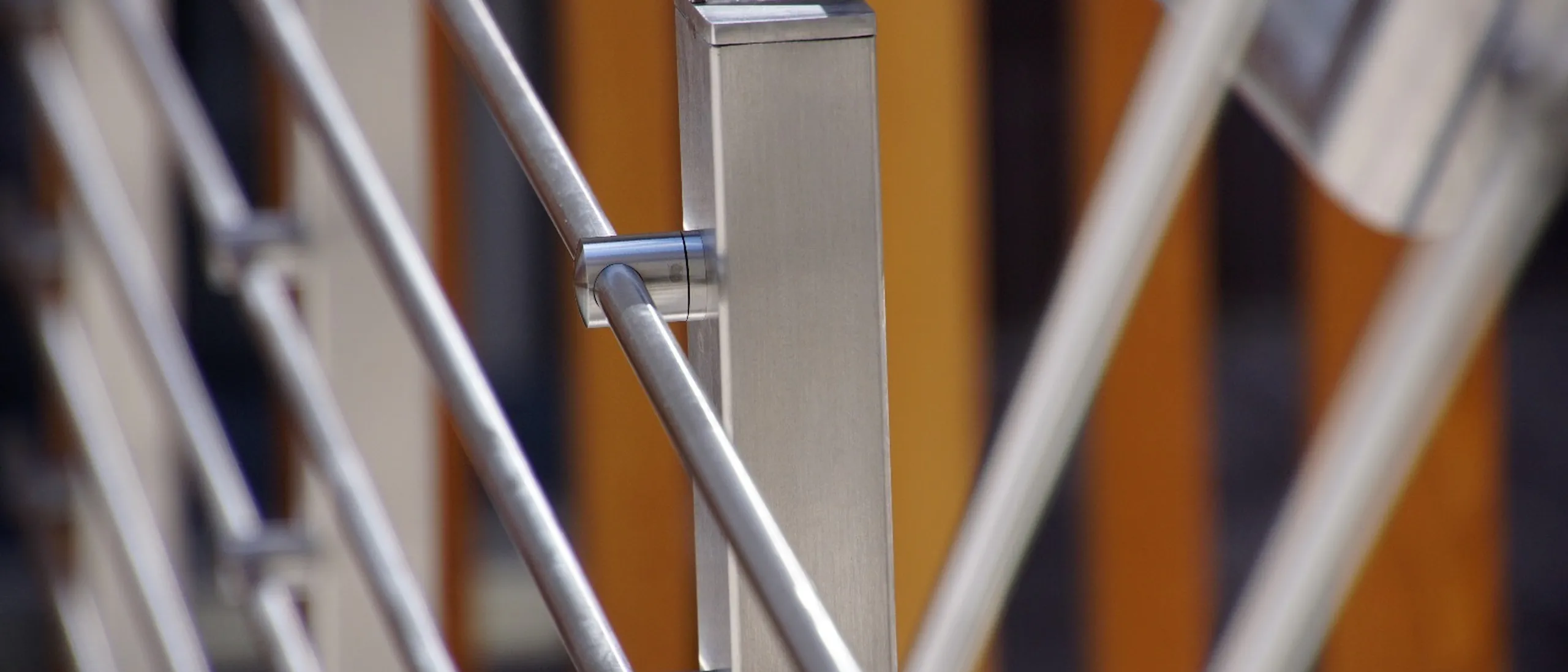 Primo piano di una ringhiera in acciaio inossidabile con barre diagonali e supporti, sfondo sfocato con elementi arancioni.