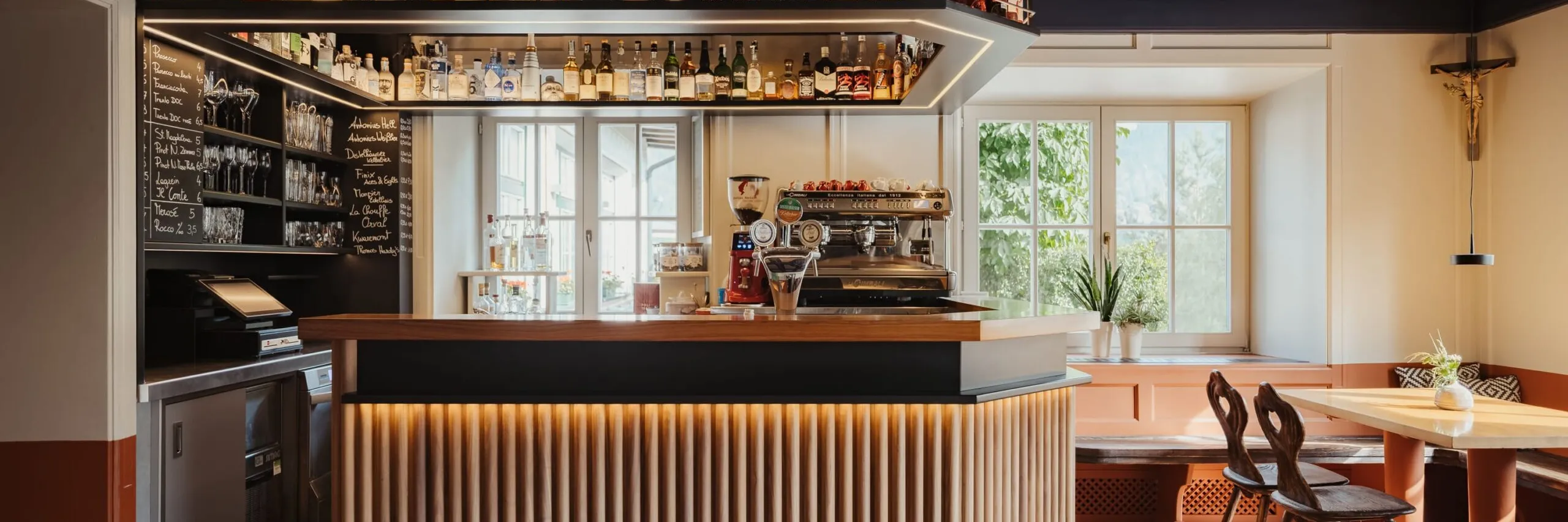 Bar moderno con bancone a listelli di legno e illuminazione a led, bottiglie esposte, macchina espresso, salotto vicino finestra, crocifisso appeso.