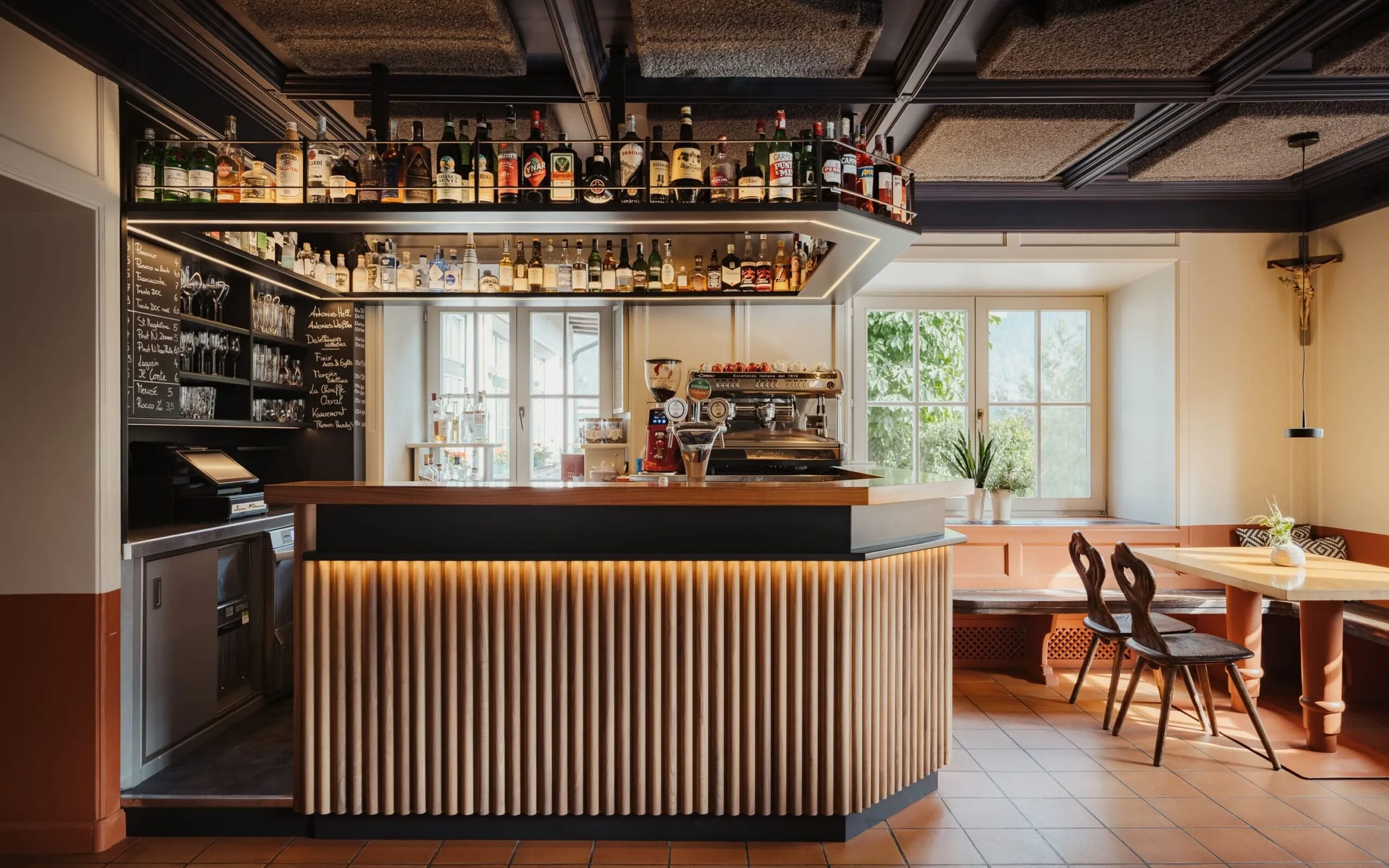Bar moderno con bancone a listelli di legno e illuminazione a led, bottiglie esposte, macchina espresso, salotto vicino finestra, crocifisso appeso.