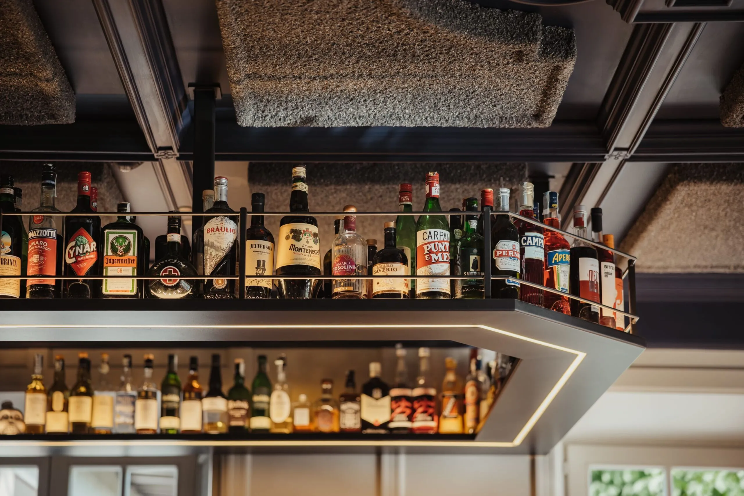 Scaffale sospeso in metallo con una fila di bottiglie di liquore su un bancone illuminato, atmosfera calda in bar.