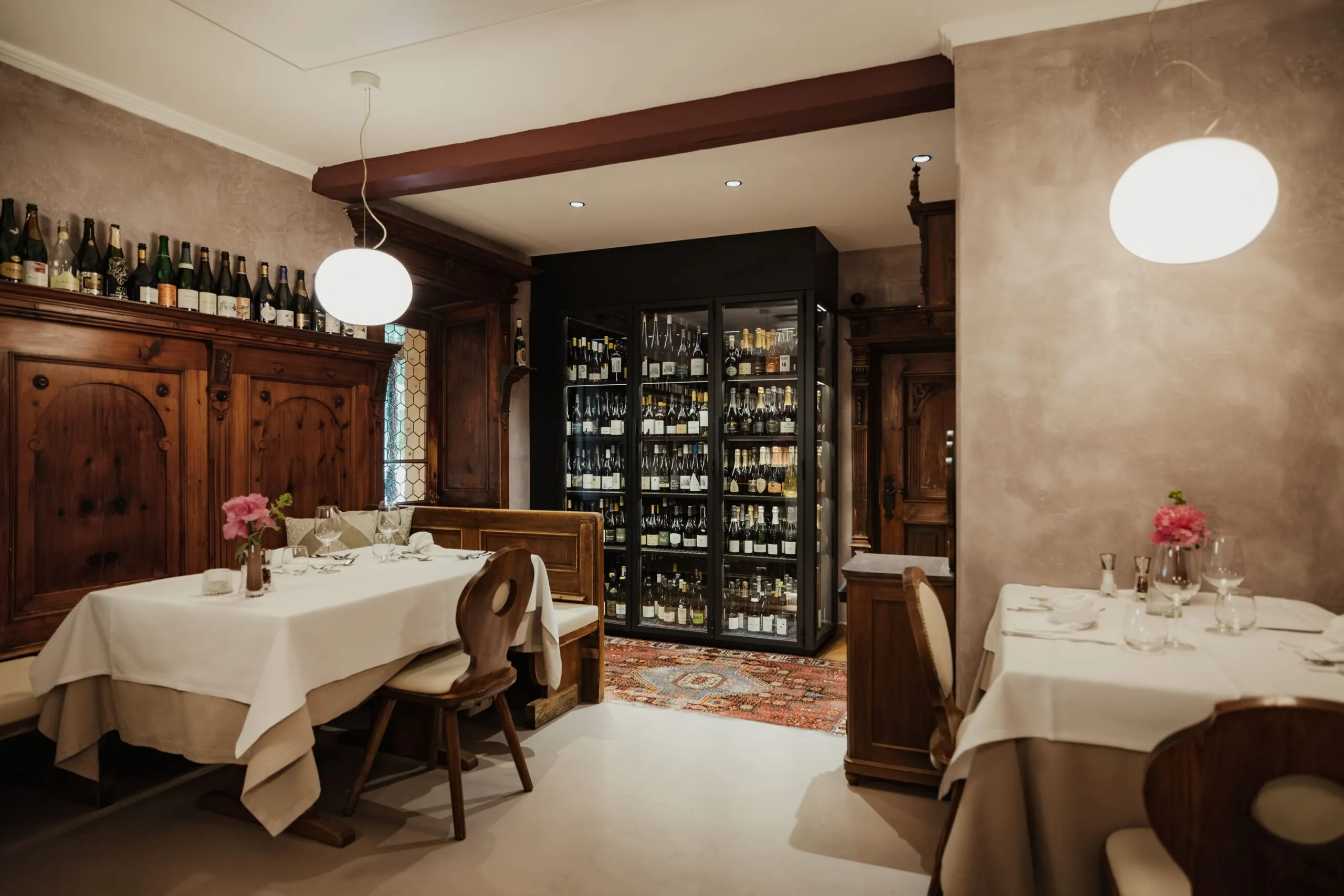 Sala ristorante con tavoli bianchi, sedie in legno, armadi in legno e una cantina di vini a vetro al centro; bouquet rosa sui tavoli, atmosfera calda.