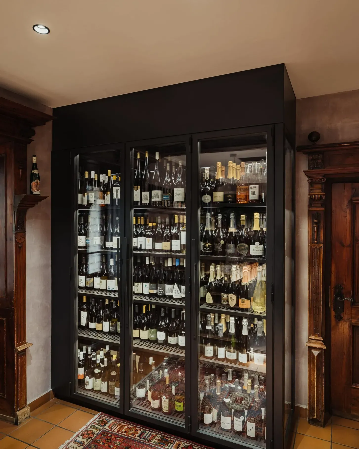 Vetrina frigorifera per vini in cornice metallica nera, ante in vetro, piena di bottiglie; sala con pavimento in cotto e arredi in legno.