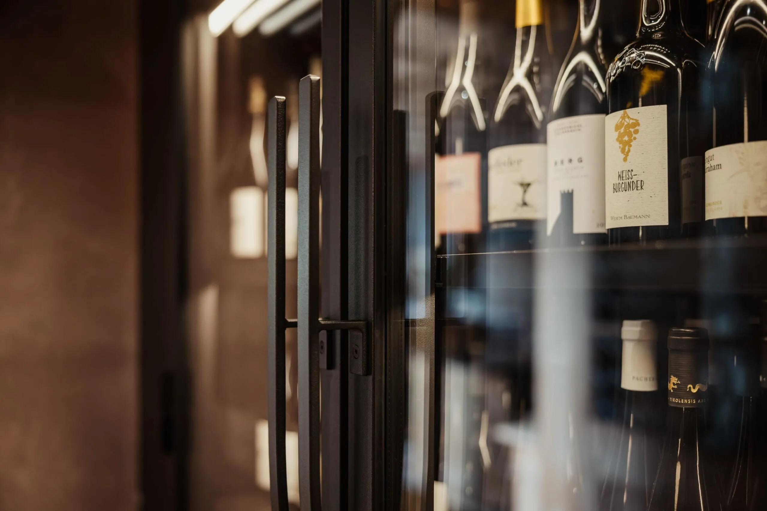 Bottiglie di vino allineate su ripiani all'interno di una cantina in vetro con cornice metallica scura; luci calde e riflessi sul vetro.
