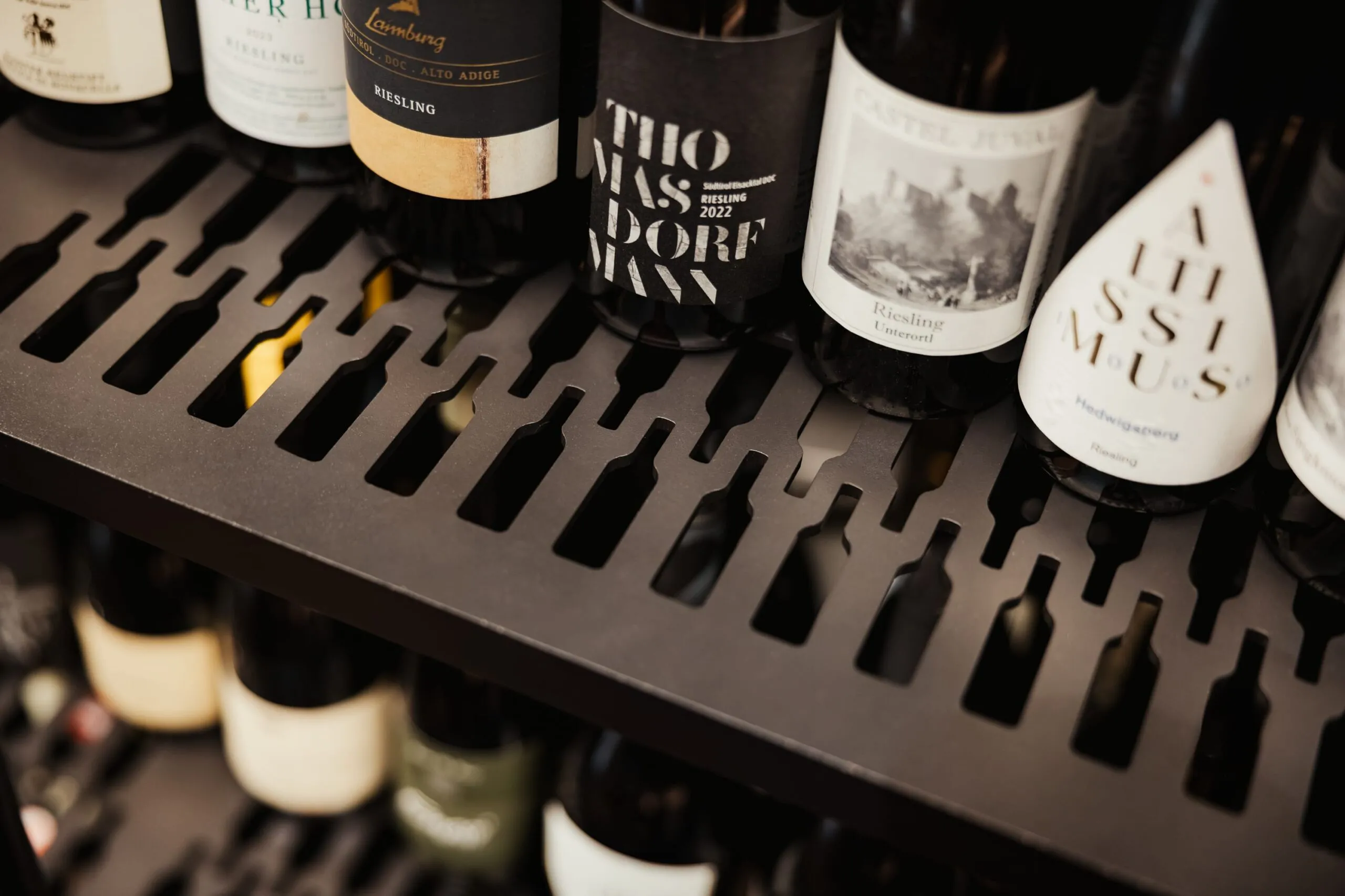 Bottiglie di vino esposte su ripiano in metallo nero con fori a forma di bottiglia; etichette visibili in una cantina/enoteca.