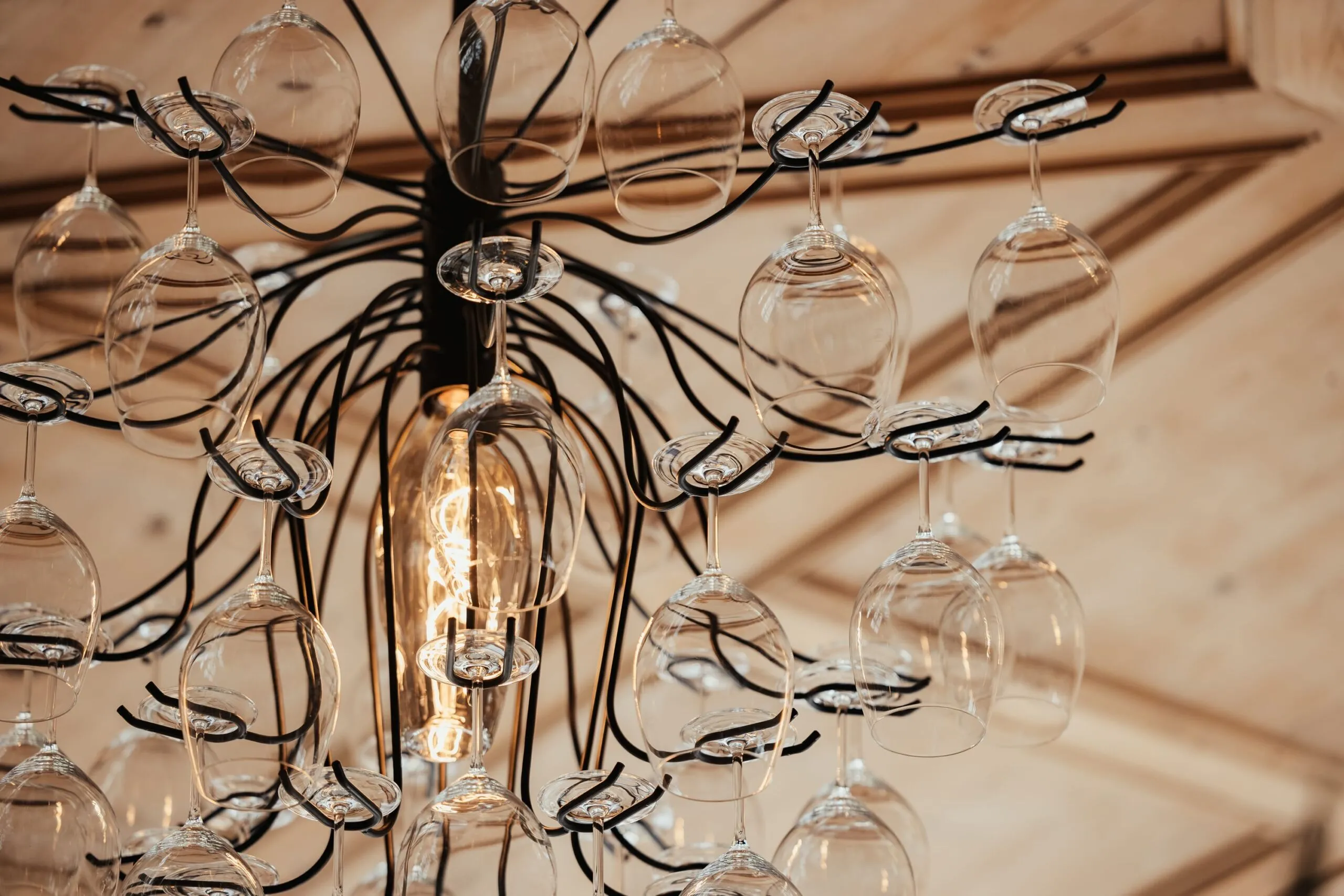 Lampadario in metallo nero con numerosi calici di vetro trasparente appesi, acceso da una lampadina centrale che emana luce calda.