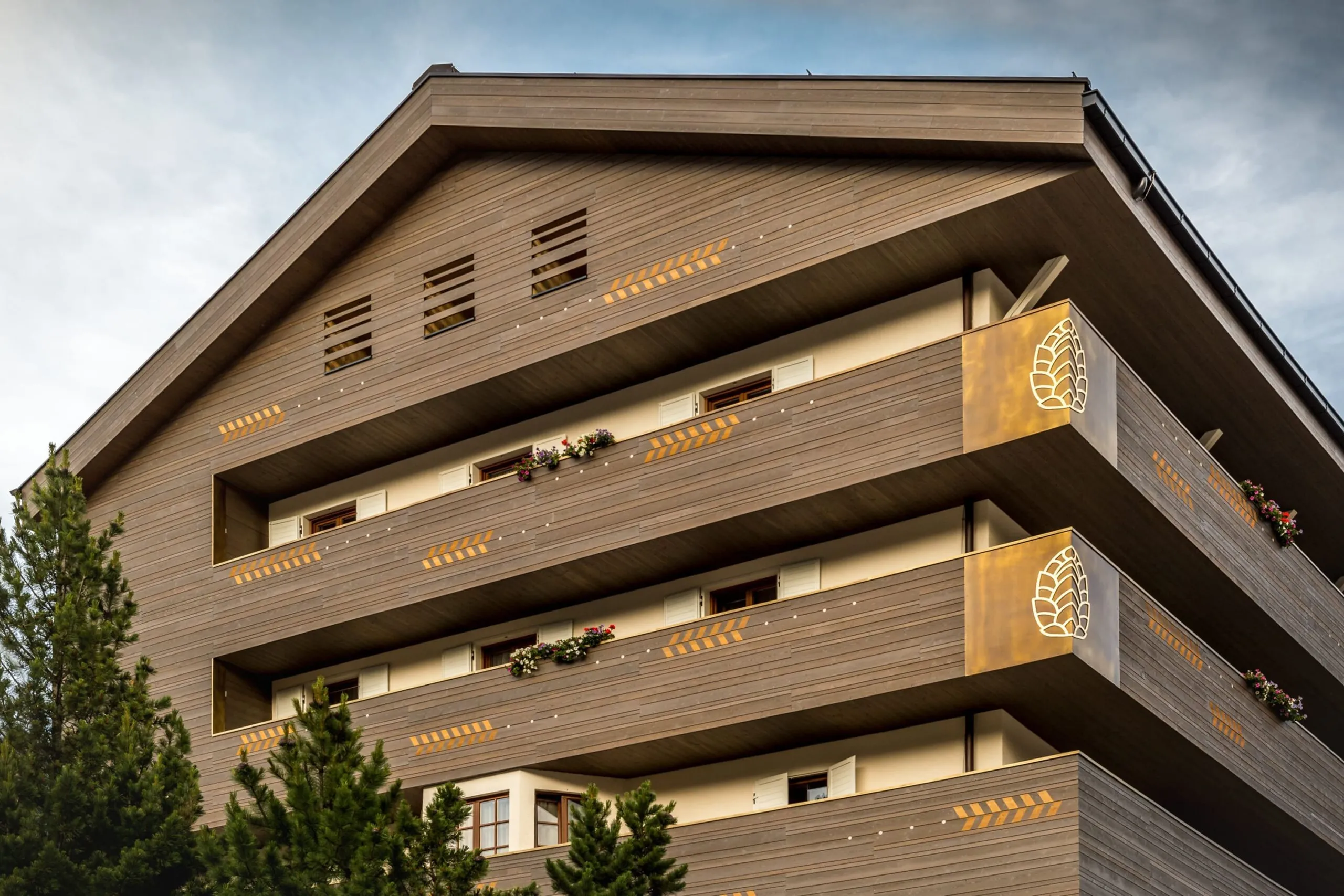 Edificio moderno in legno a più piani con balconi, fioriere e pannelli dorati con logo a forma di pigna; alberi di conifere in primo piano.