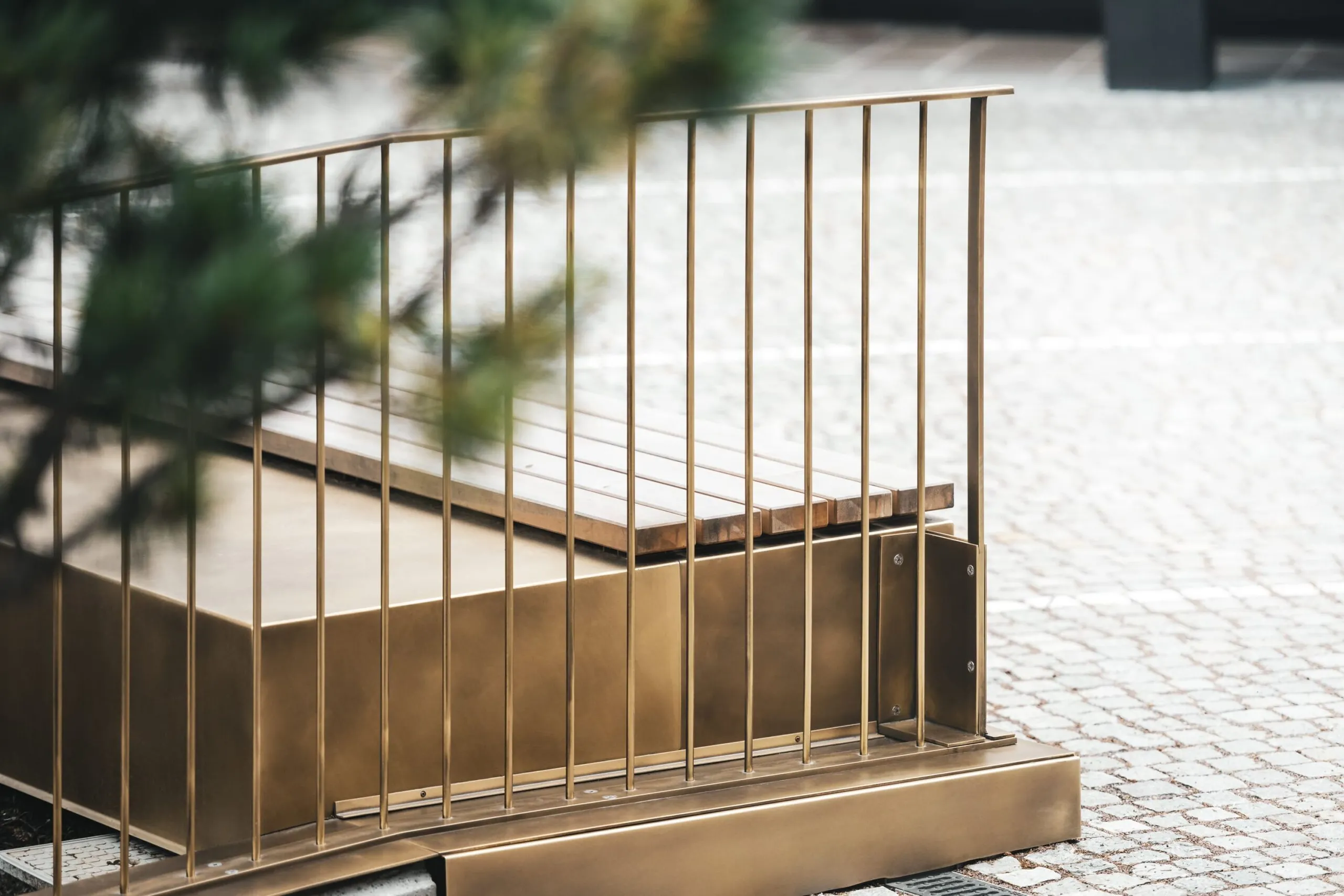 Piattaforma in metallo dorato con parapetto a barre verticali e piano in legno, su pavimentazione lastricata; sfondo sfumato.