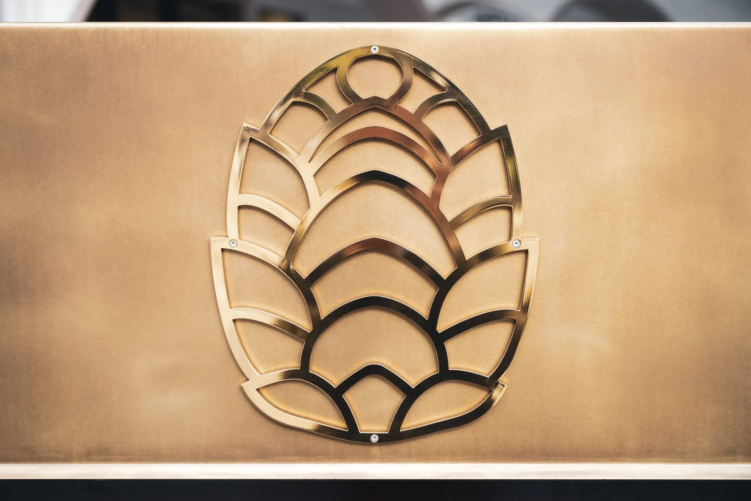 Logo aziendale in metallo a forma di foglia con archi sovrapposti, fissato su parete di tono bronzo.