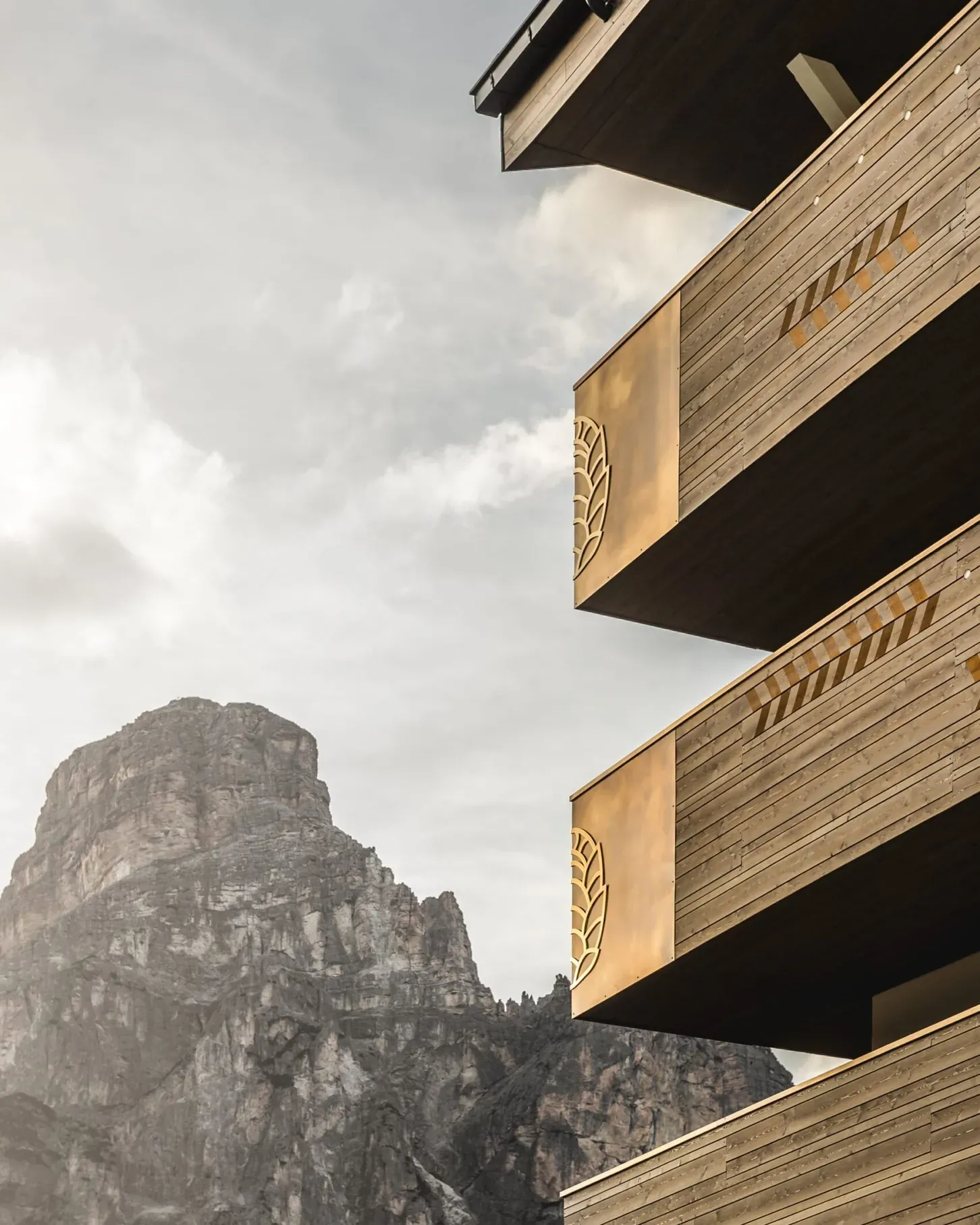 Edificio moderno in legno e metallo con balconi sovrapposti e pannelli dorati a motivo di foglia, sfondo montagna rocciosa e cielo nuvoloso.