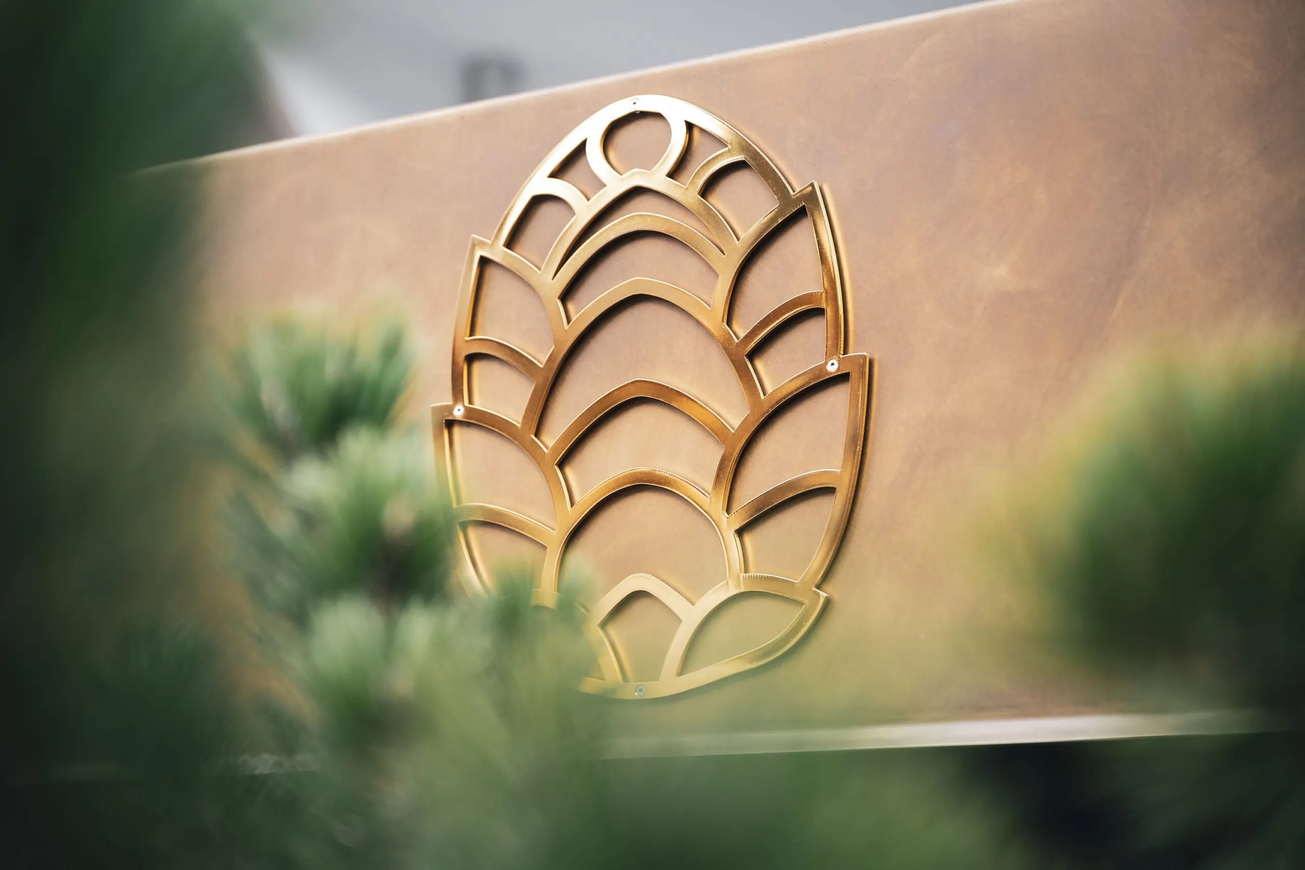 Emblema metallico dorato a forma di foglia stilizzata con motivi ad arco, fissato su una parete metallica; piante verdi sfocate in primo piano.