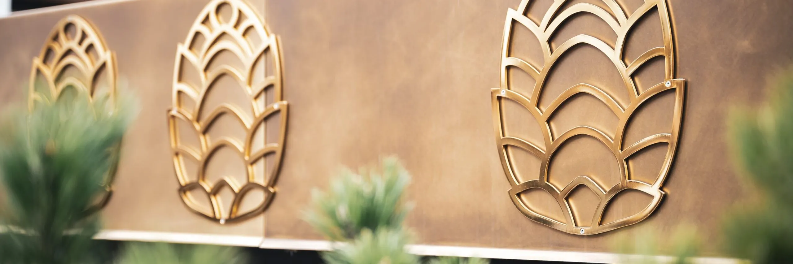 Pannello metallico marrone-dorato con tre motivi stilizzati a foglia in ottone; piante verdi sfocate in primo piano.