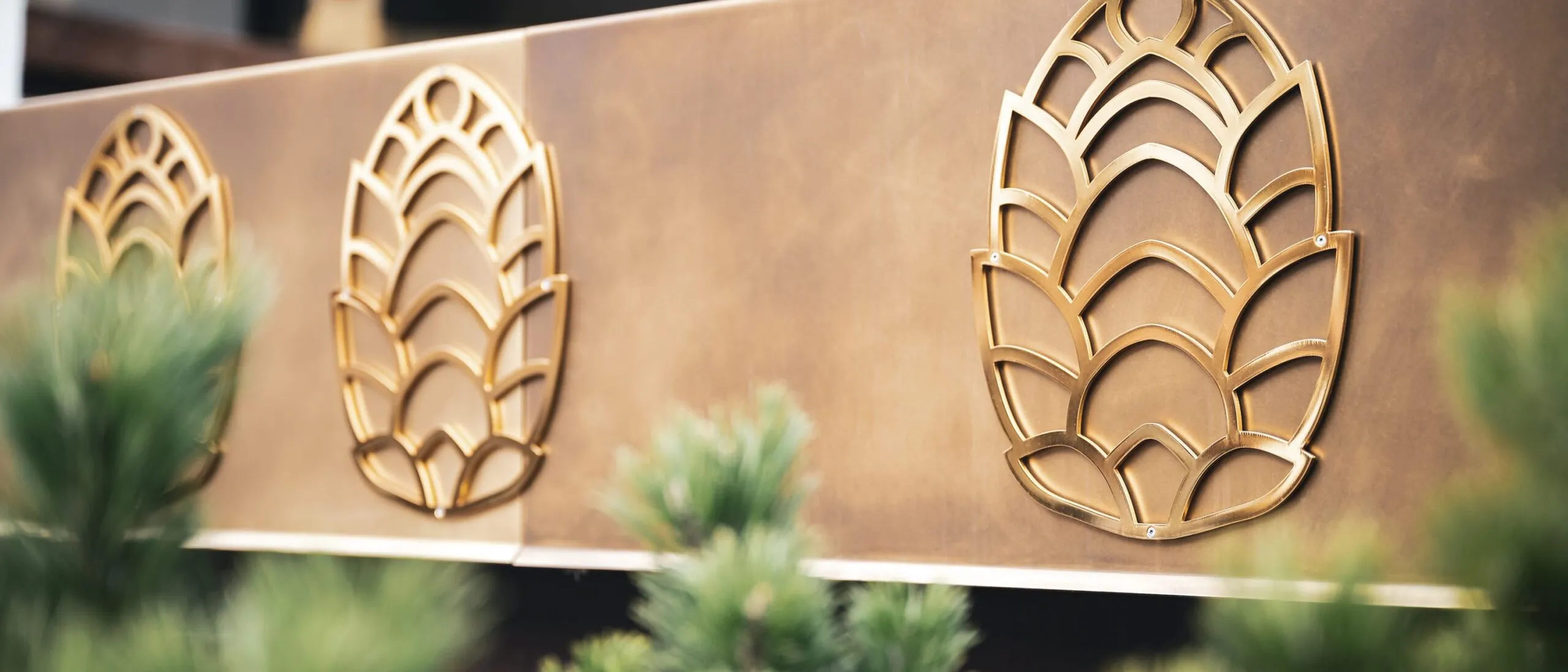 Pannello metallico marrone-dorato con tre motivi stilizzati a foglia in ottone; piante verdi sfocate in primo piano.