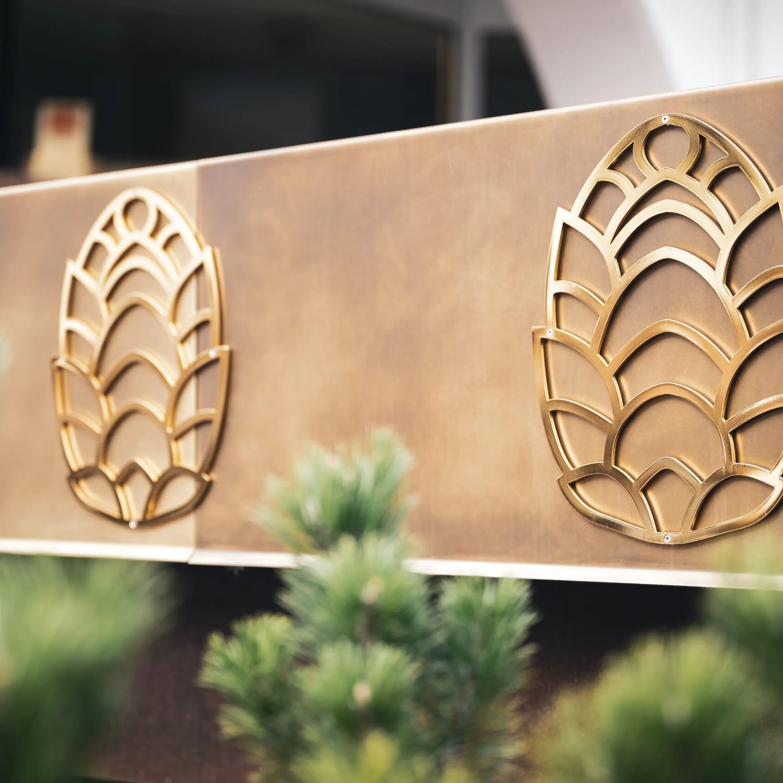 Pannello metallico marrone-dorato con tre motivi stilizzati a foglia in ottone; piante verdi sfocate in primo piano.