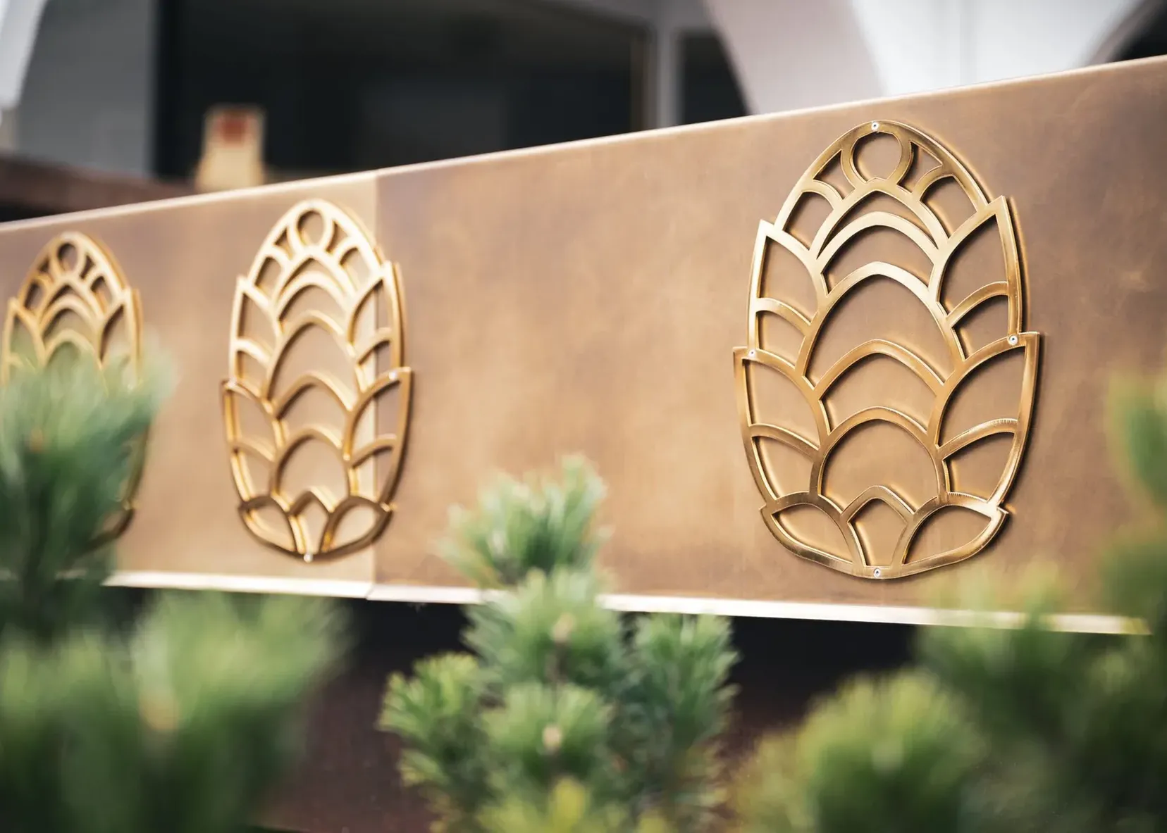Pannello metallico marrone-dorato con tre motivi stilizzati a foglia in ottone; piante verdi sfocate in primo piano.