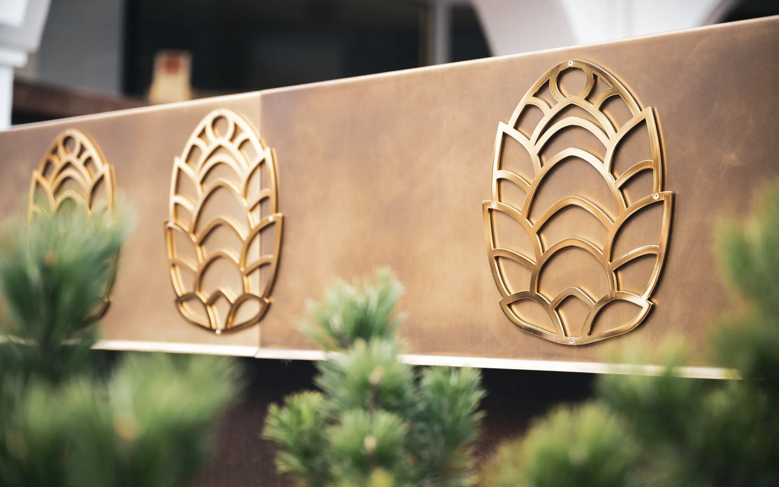Pannello metallico marrone-dorato con tre motivi stilizzati a foglia in ottone; piante verdi sfocate in primo piano.