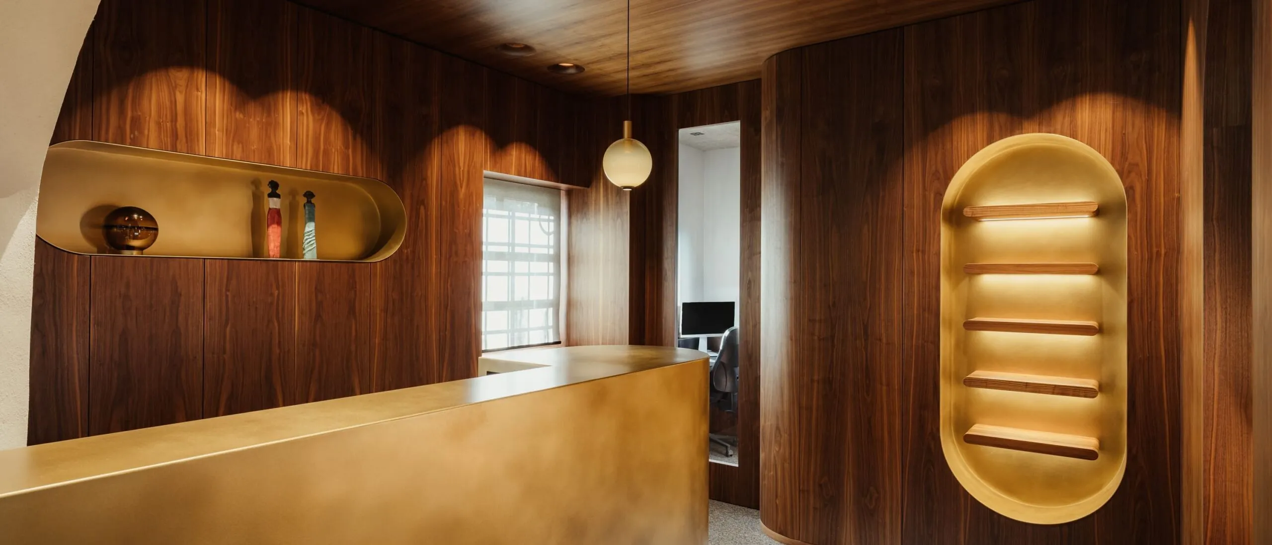 Interno di un'azienda metalmeccanica in Alto Adige: pareti e soffitto in legno scuro, reception dorata, nicchie illuminate e zona lavoro con computer.