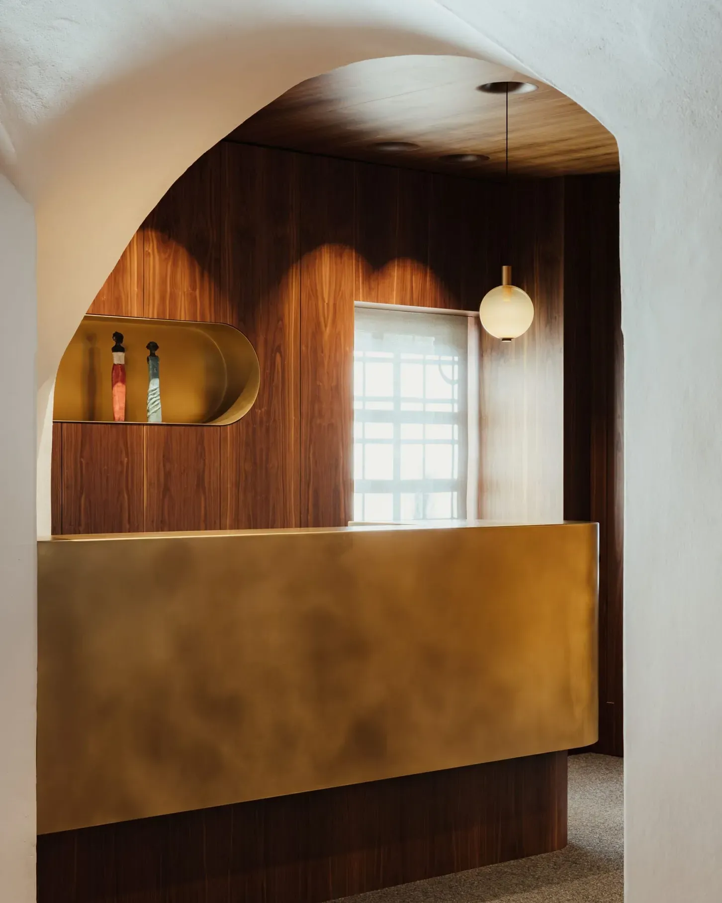 Interno reception in legno scuro con bancone dorato curvo, arco bianco, lampada a sfera e nicchia con due figurine; finestra a griglia sullo sfondo.