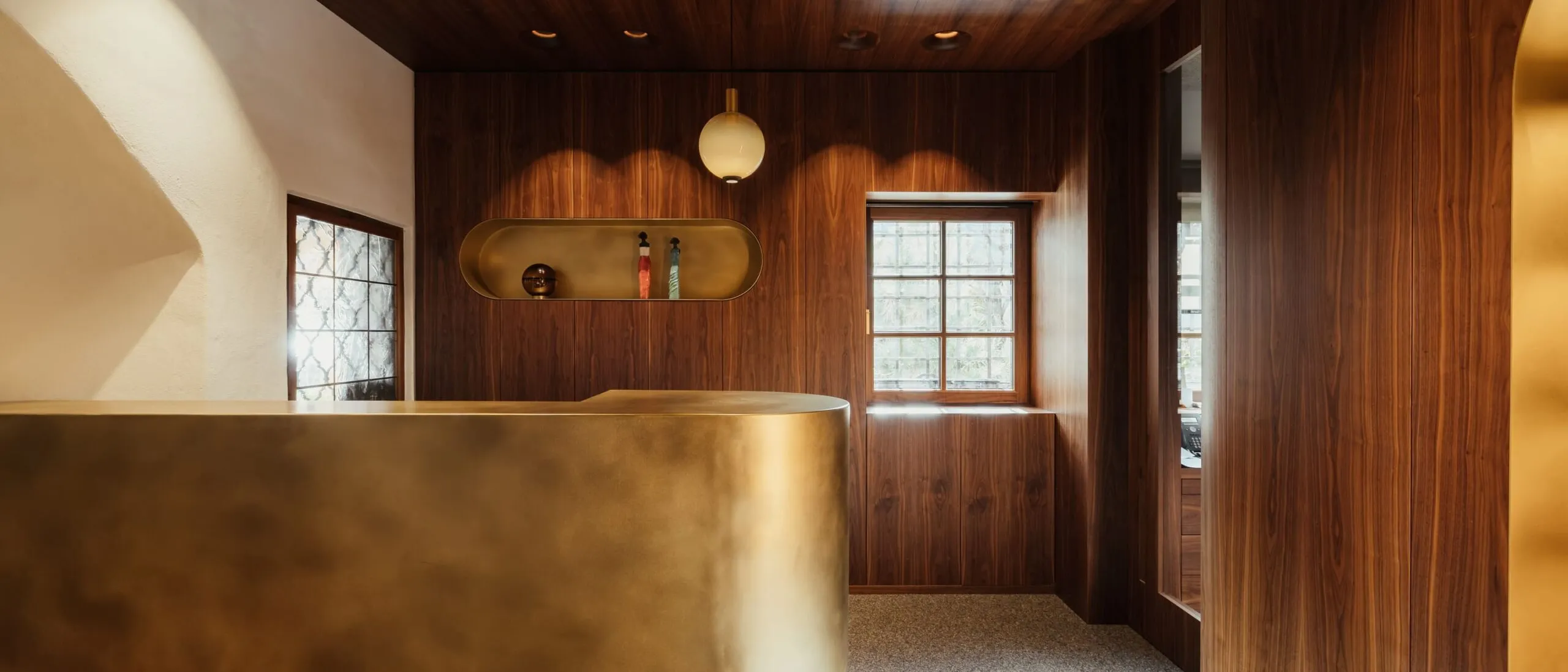 Reception in legno scuro con banco dorato curvo, mensola ovale incassata con bottiglie, finestra a grate, luci soffuse e pareti rivestite in legno.