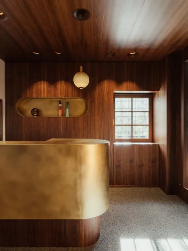 Reception in legno scuro con banco dorato curvo, mensola ovale incassata con bottiglie, finestra a grate, luci soffuse e pareti rivestite in legno.