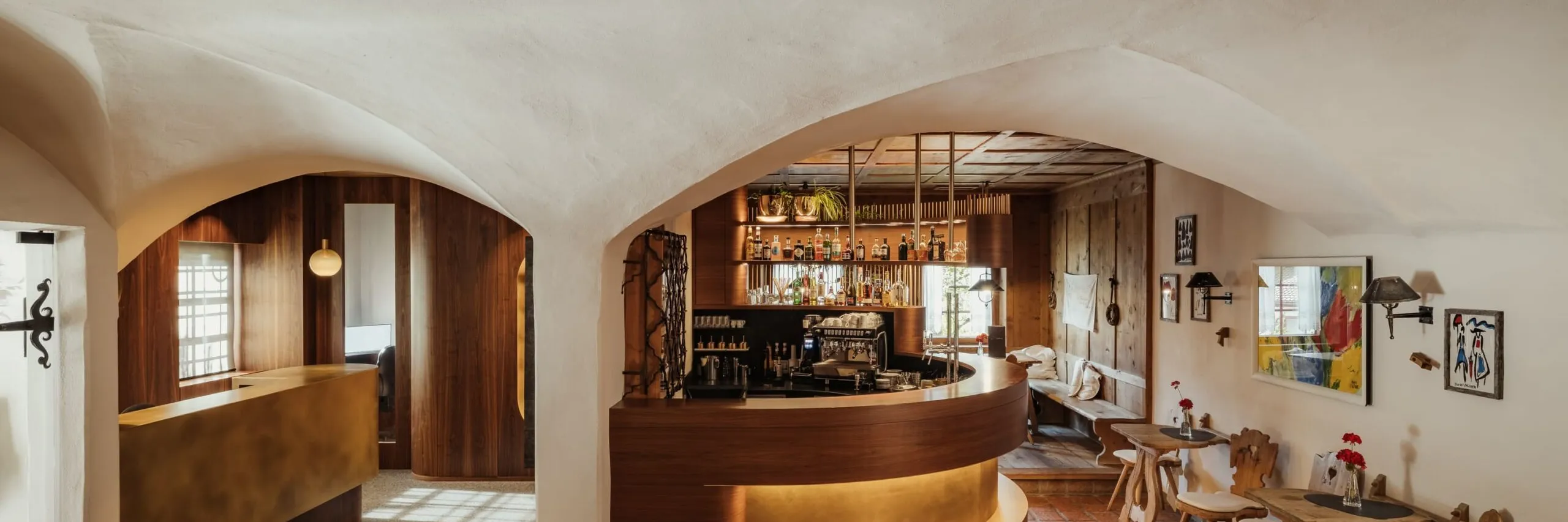 Interno di un bar ristorante con soffitti in legno a travi, archi bianchi, grande bancone curvo in legno e luci calde; tavolini e sedie lungo la parete.