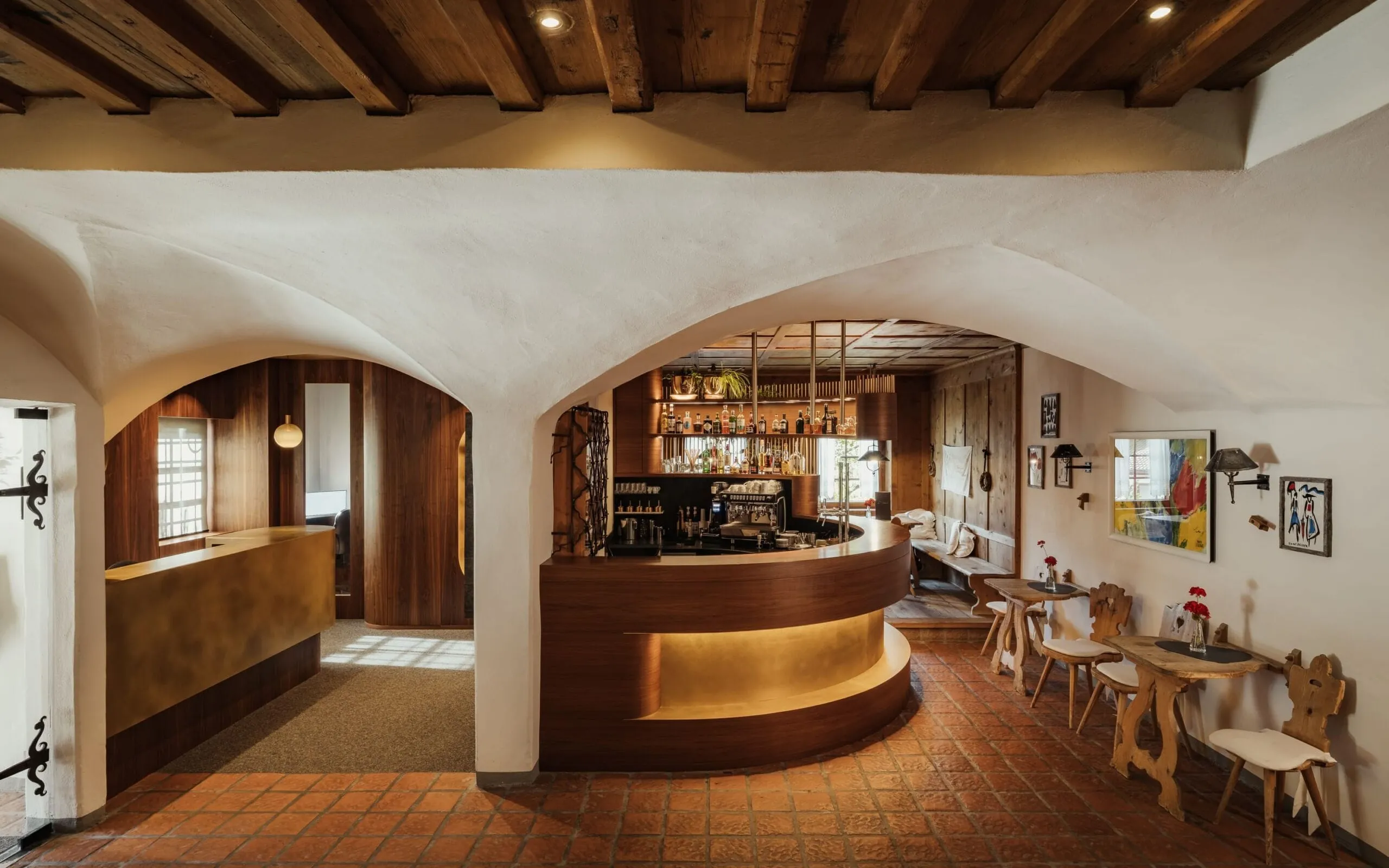 Interno di un bar ristorante con soffitti in legno a travi, archi bianchi, grande bancone curvo in legno e luci calde; tavolini e sedie lungo la parete.