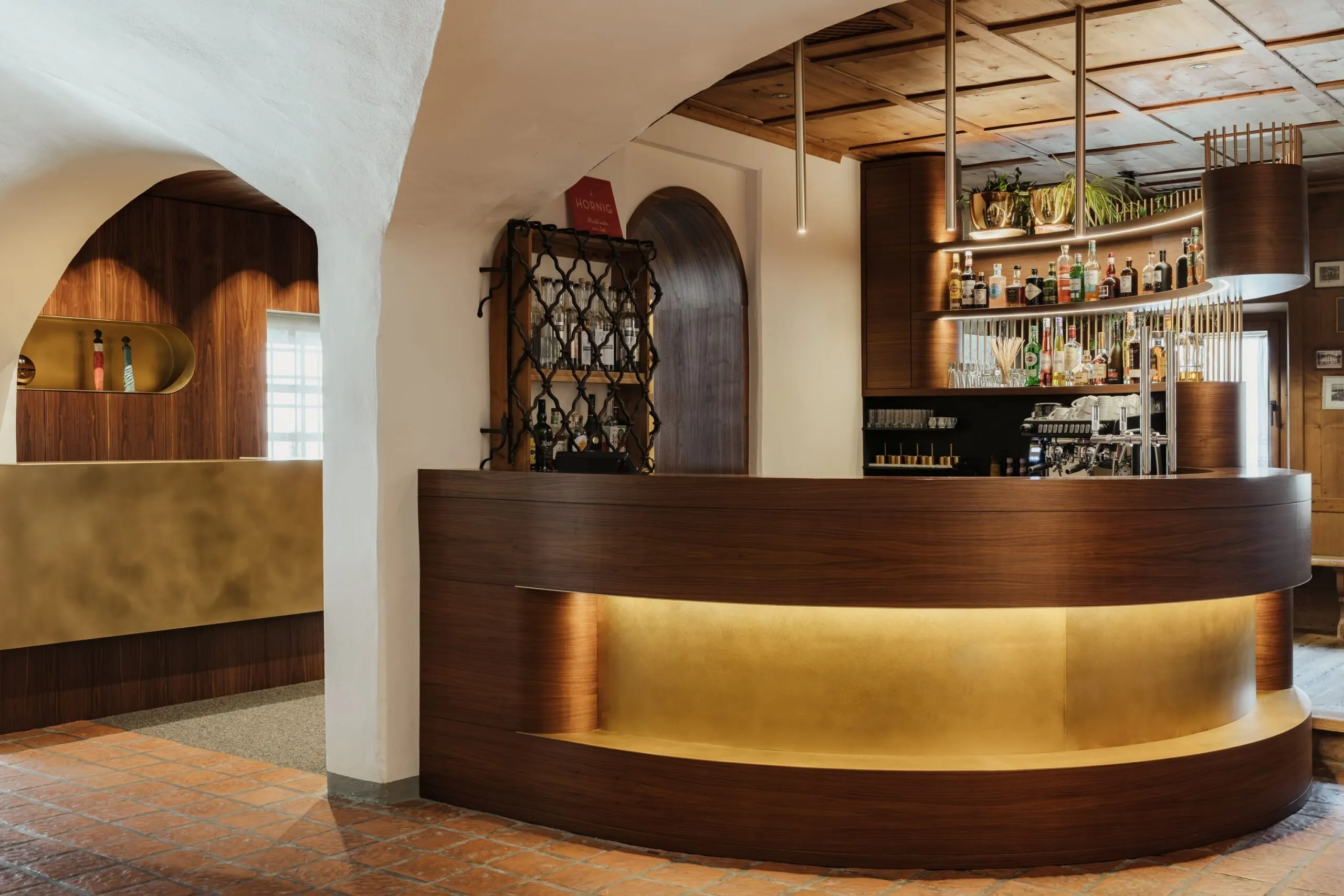 Interno bar in legno con bancone semicircolare illuminato, retrobar con bottiglie e bicchieri, scaffale vino, archi e pavimento in cotto.