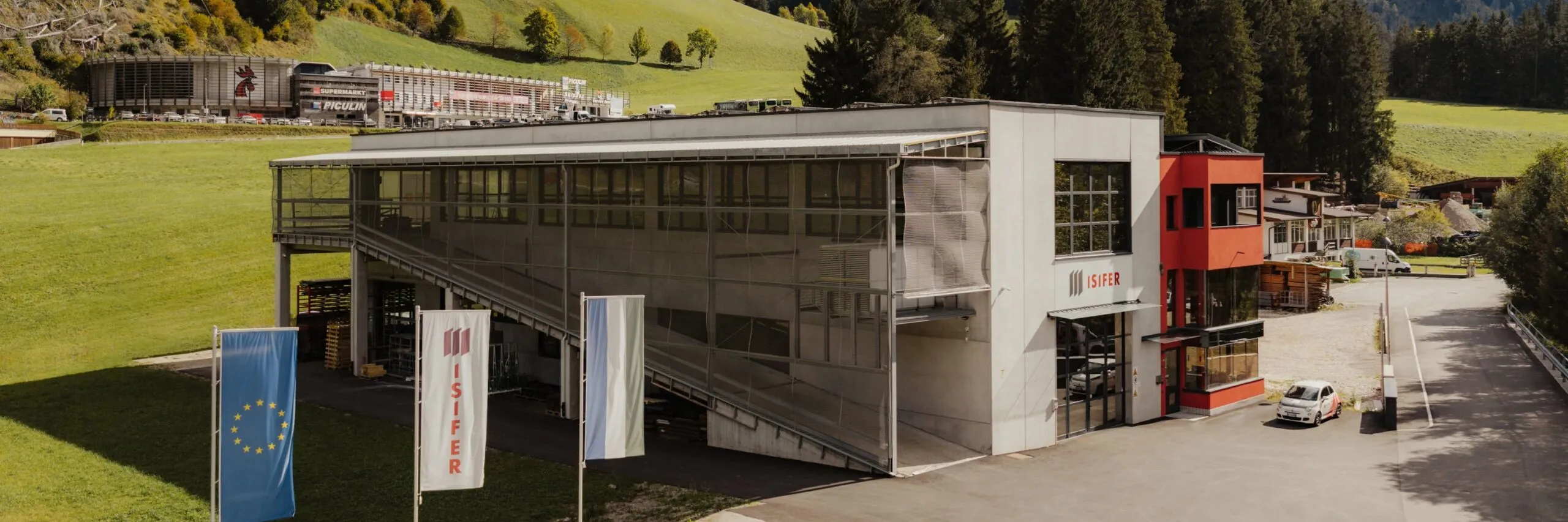 Außenansicht eines modernen Metallbau-Standorts in Südtirol mit Glasfront, roter Akzentwand, EU-Flagge und ISIFER-Flaggen; grüne Hänge und Berge im Hintergrund.