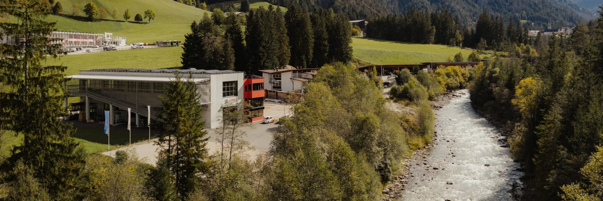 Landschaft mit Fluss, Wald und Wiesen; modernes Metallbau-Unternehmen am Ufer, Südtirol, Bergkulisse und blauer Himmel.