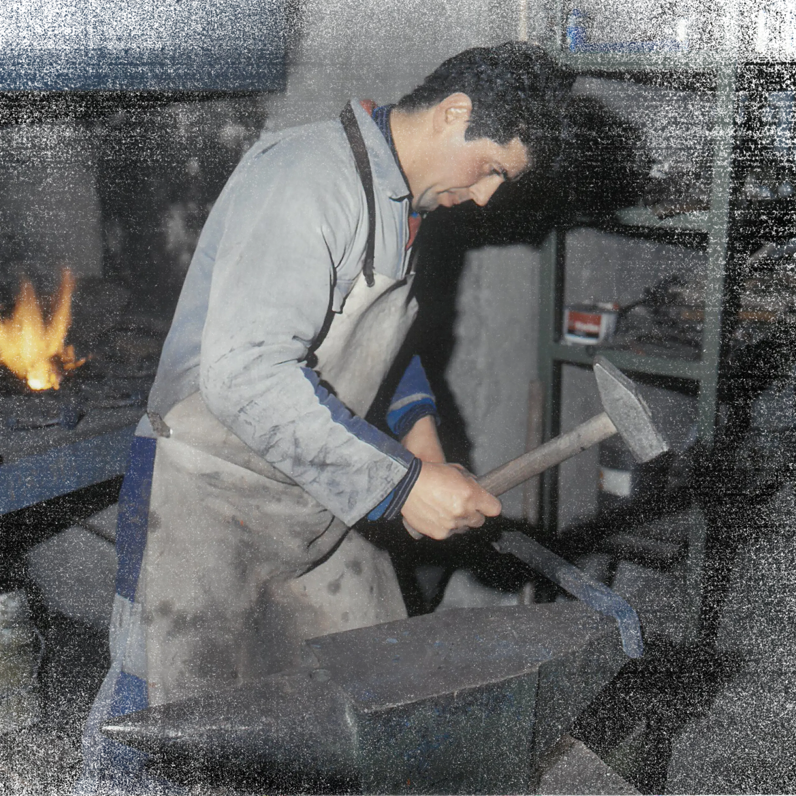 Schmiedende Person in einer Südtiroler Metallbau-Werkstatt, arbeitet am Amboss mit Hammer in der Hand; Flamme im Hintergrund, Schürze getragen.